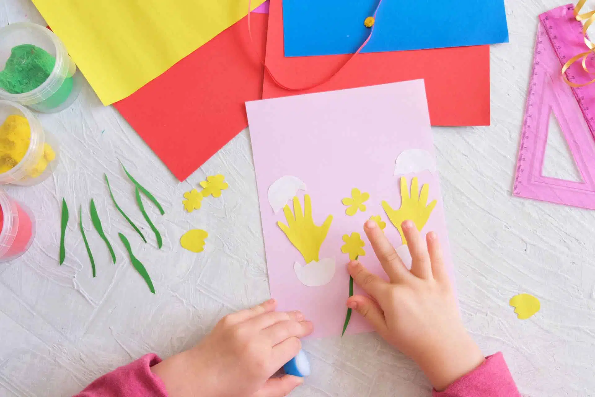 On voit les mains d'un enfant qui effectue du bricolage. On voit des feuilles de couleurs rose, rouge, bleue et jaune ainsi que des pots de pâtes à modeler et une règle.