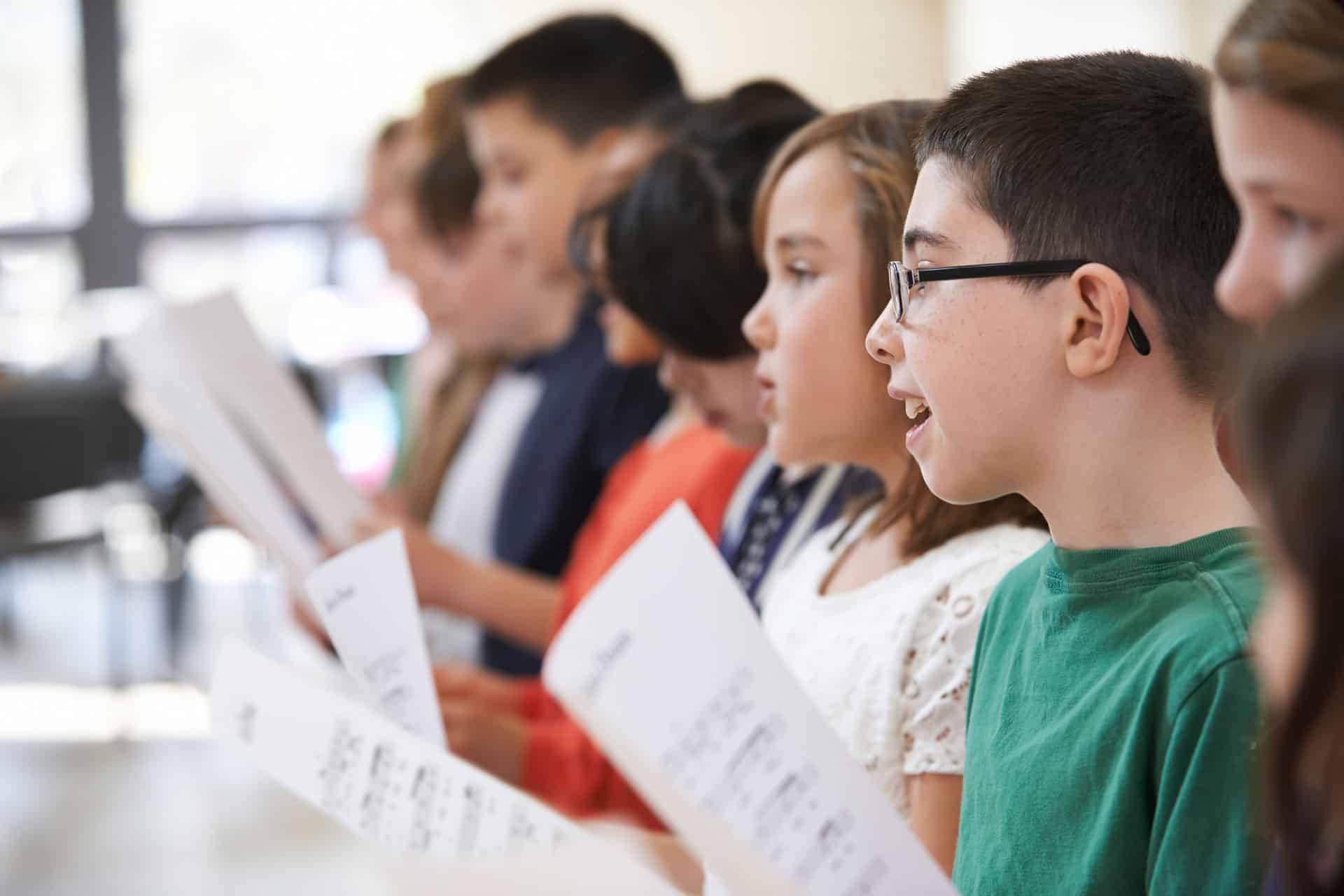 Un groupe de 7 enfants chantent dans une chorale avec des feuilles de partition dans leurs mains.