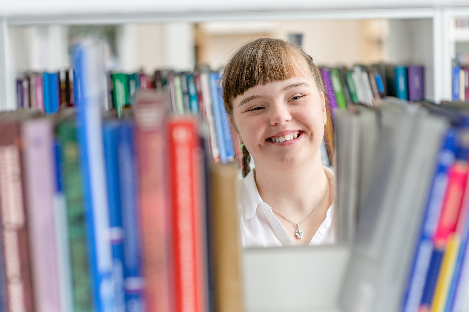 Jeune fille souriante que l'on voit au travers des livres placés sur une étagère de bibliothèque.